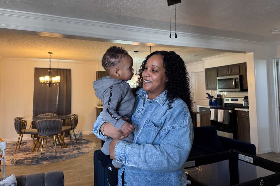 Holding her toddler child, Kennisha Missouri stands in the dining area of her home in Petersburg, Virginia. A kitchen is in the background.