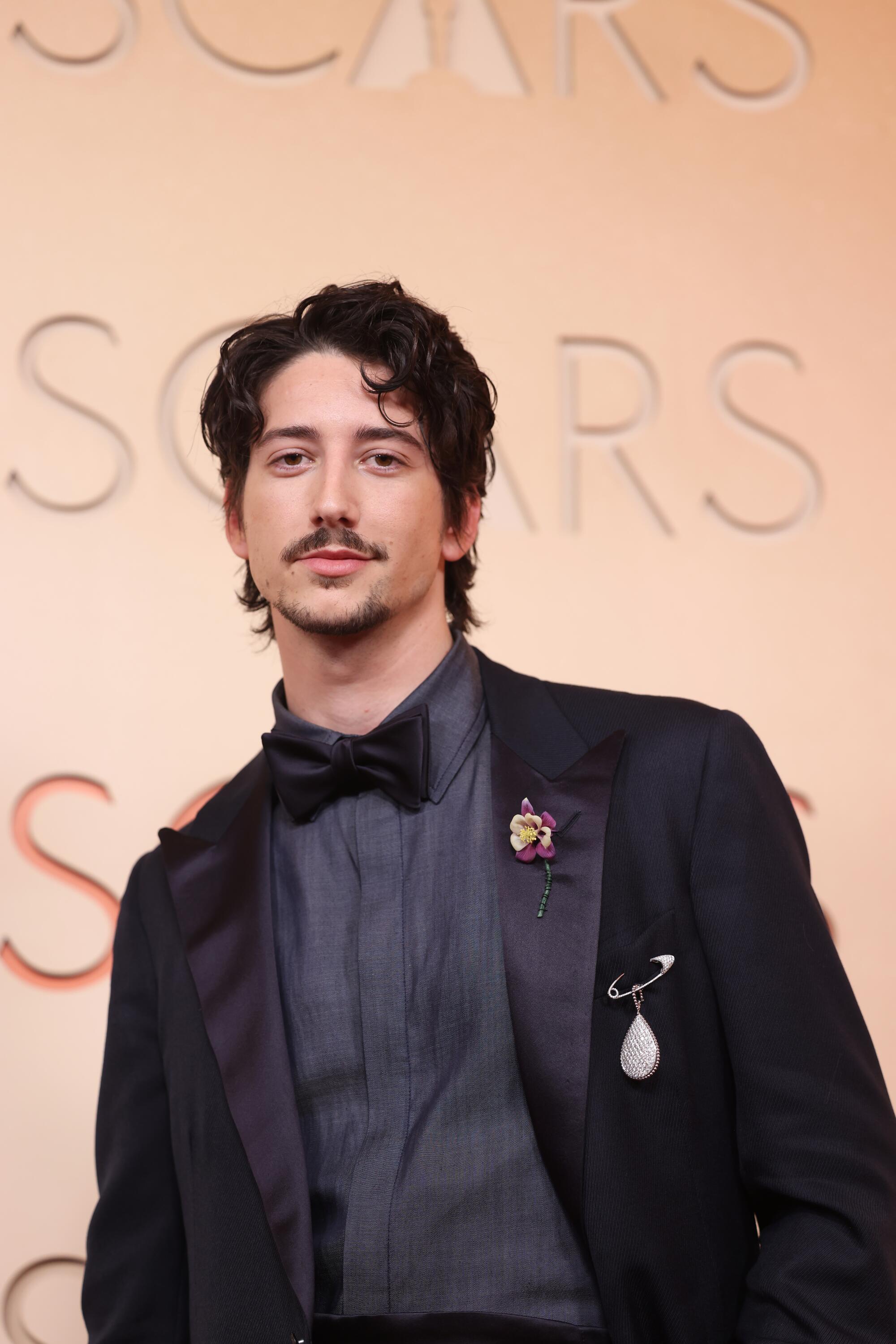 Milo Manheim, wearing an all-black tux, poses on the red carpet. 