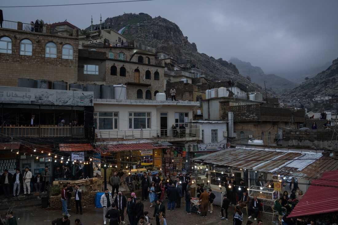 Dusk falls over Akre on Nowruz, a celebration of spring and renewal, and people gather in a central plaza. Mountains and homes loom over the plaza, which has twinkle lights strung up.