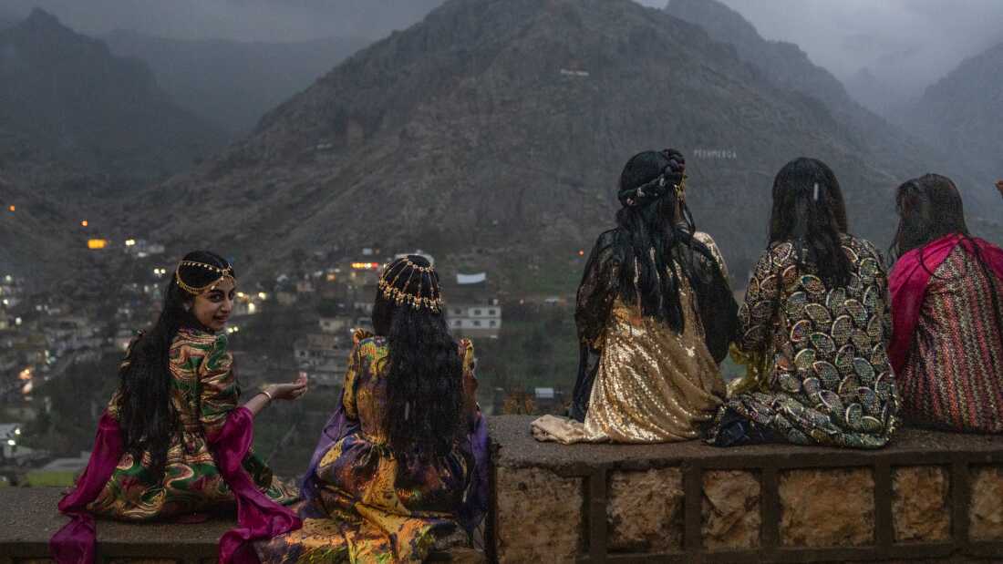 Five teen girls sit on a low wall to get a good vantage point of people walking up the mountains with flaming torches and fireworks. They are wearing colorful gowns and some have gold headpieces over their long hair.