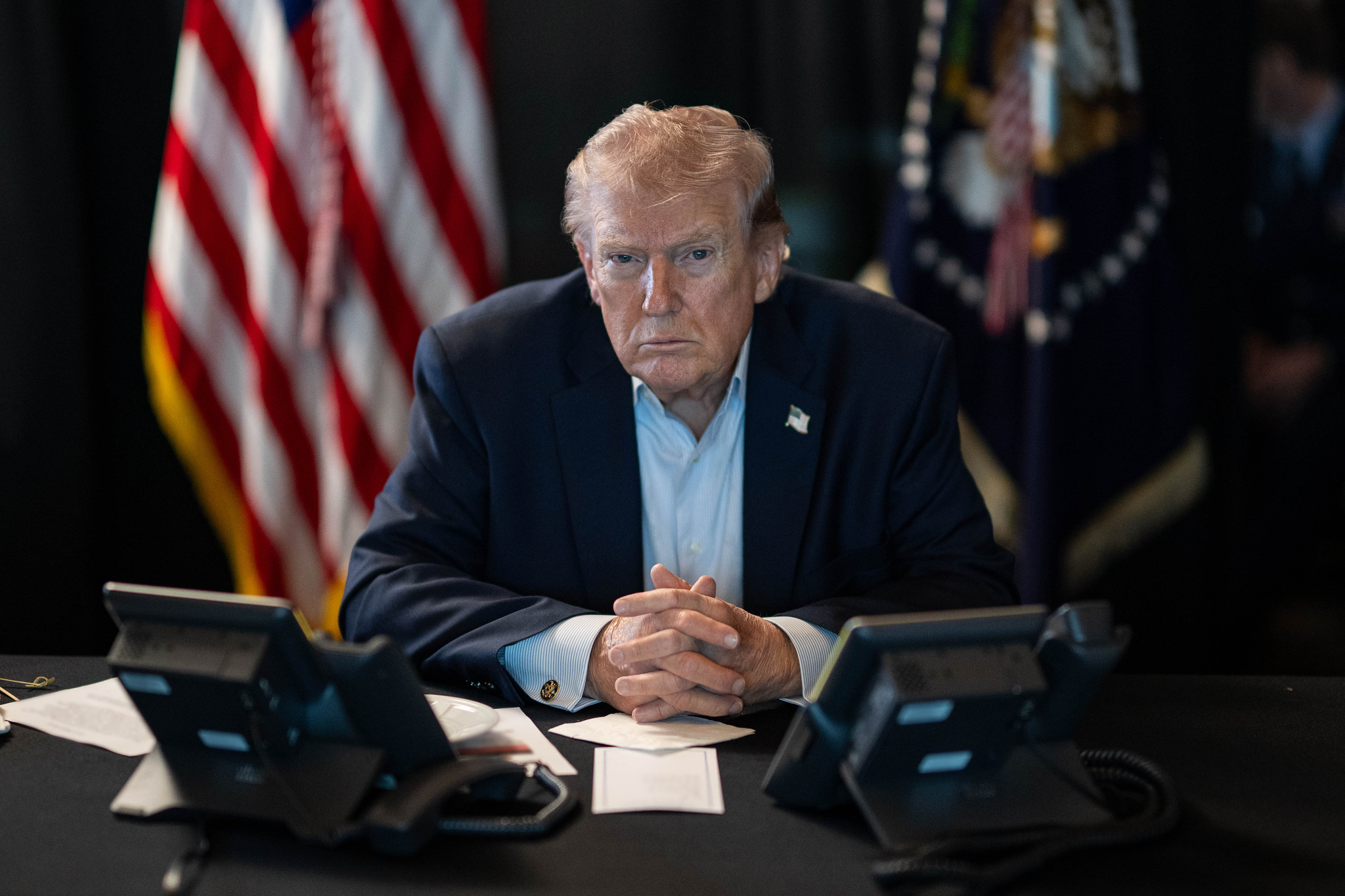 Donald Trump seated at a desk with two phones and an American flag in the background.
