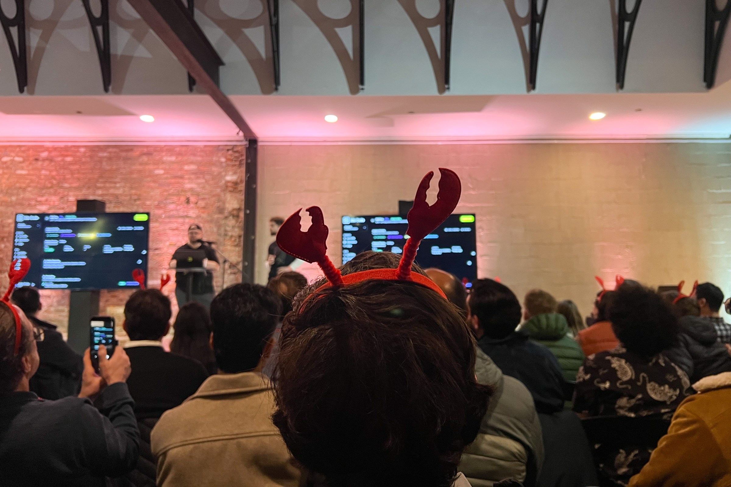 A man in a lobster claw headband, seen from the back, looking out on a crowd of people and a stage.