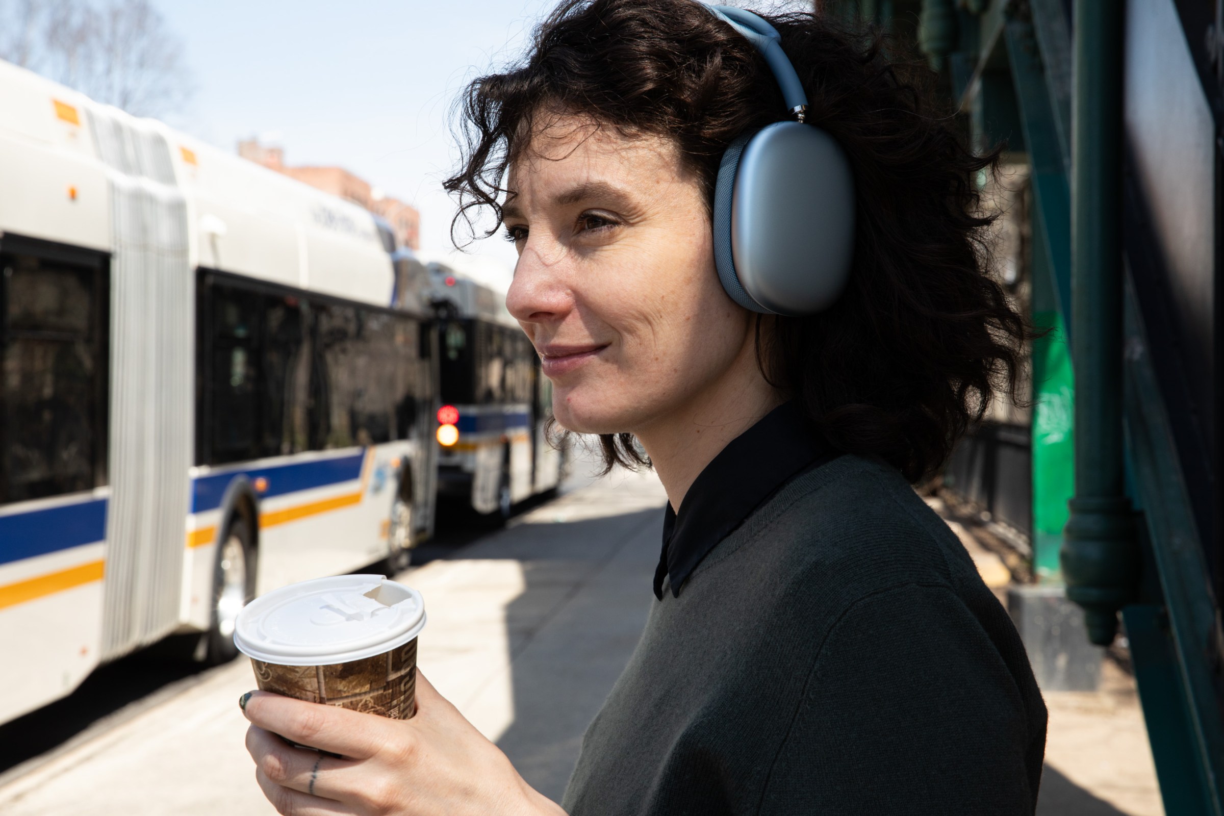 A person wearing the blue Apple AirPods Max 2 while holding a coffee outside.