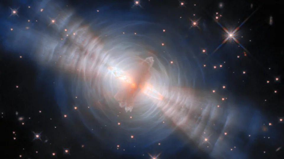 An image of the egg nebula, with a glowing streak of pink gas in the middle surrounded by concentric circles of white like with four diagonal beams of light streaking from top left to bottom right all in front of a deep space starry background