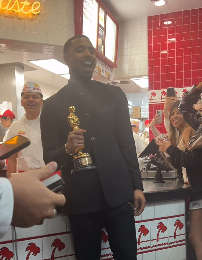 Michael B. Jordan holding an Oscar trophy inside a fast-food restaurant surrounded by fans taking photos.