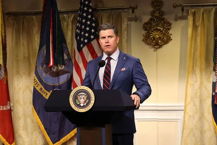 Person in a suit speaks at a podium with a presidential seal, flanked by flags. Background features decorative elements