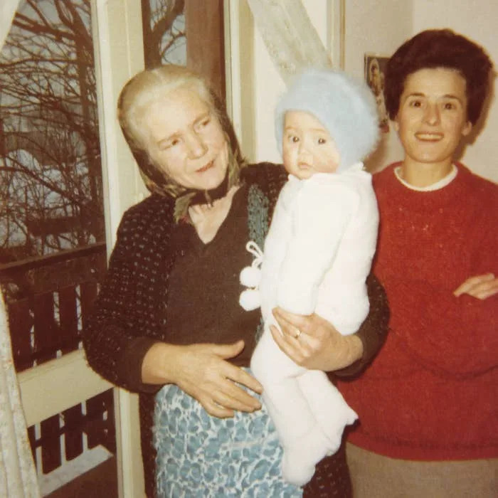 Two women stand indoors by a window. One holds a baby dressed in a fluffy outfit and beanie