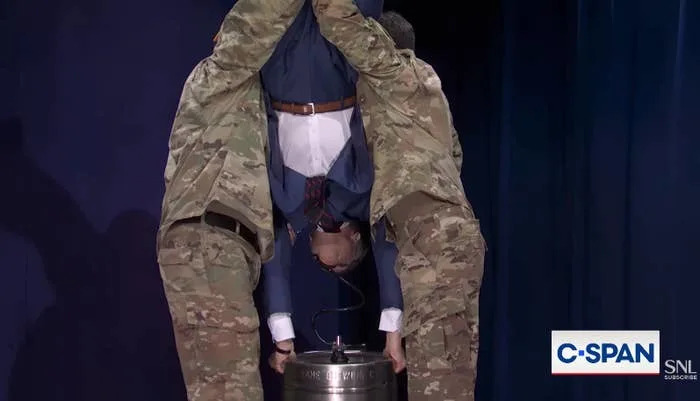 A man in a suit is held upside down over a keg by two people in military uniforms during a comedic skit