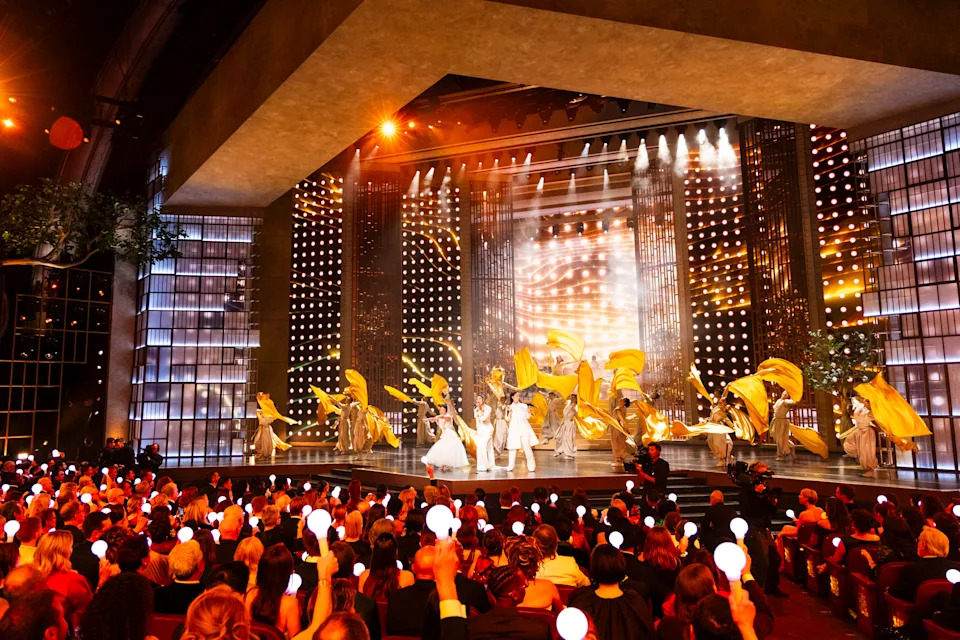 Wide view of a theater stage performance with dancers waving large gold fabric flags while the seated audience holds glowing lights.