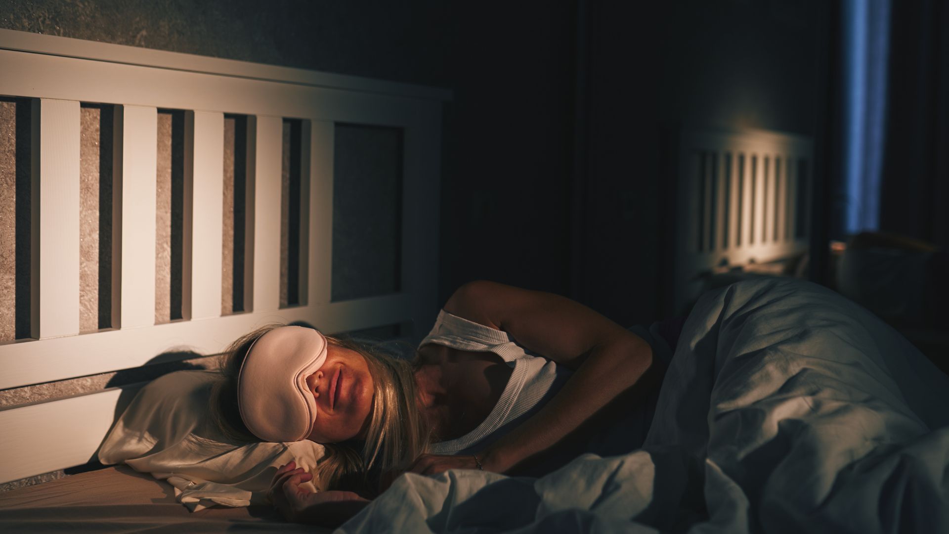 A woman sleeps happily in bed at night - she is lying on her side with a white eye mask on.