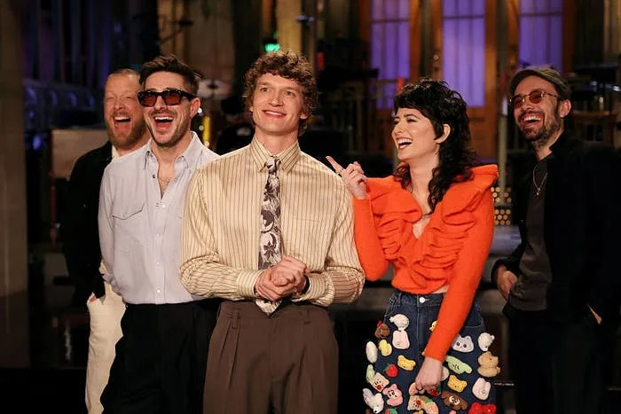 Group of five people laughing together indoors, one wearing a ruffled top with cartoon patches. They're on a studio set, possibly for a TV show