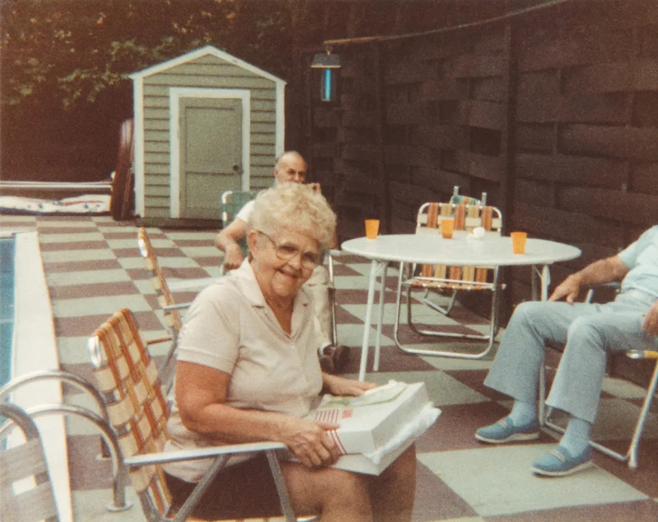 An elderly woman sits by a poolside patio table reading a book, while two others relax on chairs nearby