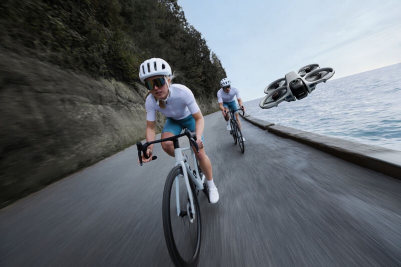 Two cyclists wearing white helmets and blue shorts ride along a lakeside road, while a drone flies nearby capturing footage. Trees line the left side, and calm water is on the right.