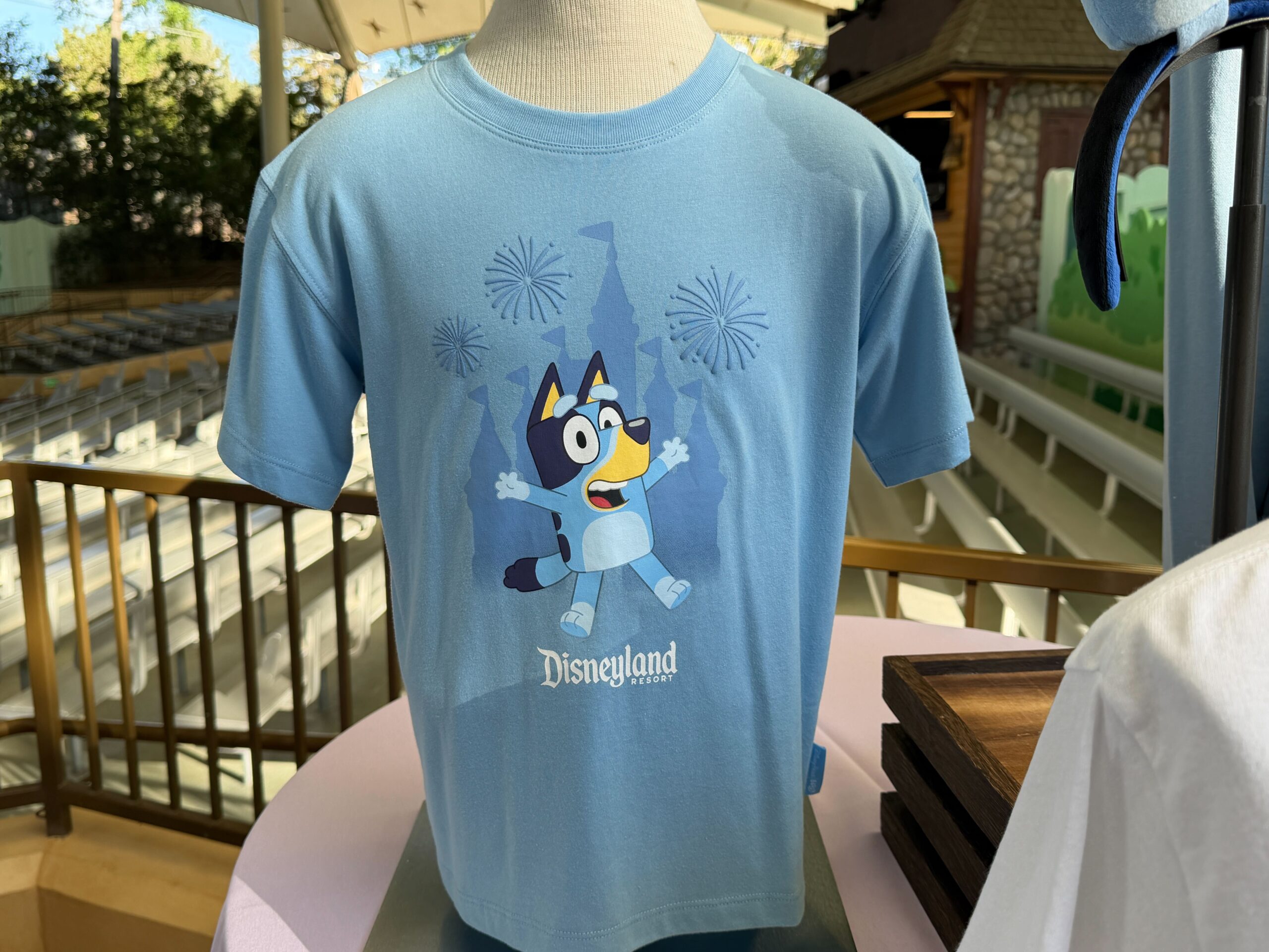 On the left edge of the display, we have a light blue short-sleeved graphic T-shirt. The torso graphic features Bluey in a cheerful pose in front of a Sleeping Beauty castle silhouette, with small fireworks bursting around her. “Disneyland Resort” is printed near the bottom of the design in a dark blue font.