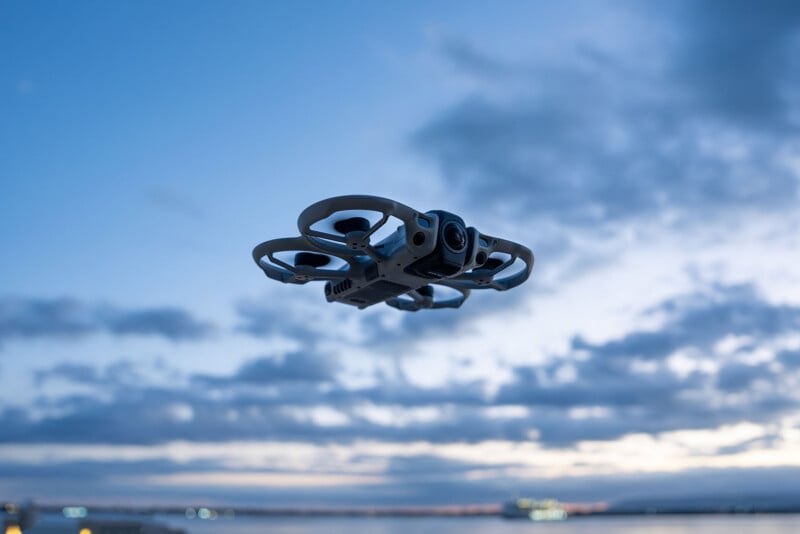 A small drone with protective propeller guards hovers in the air against a backdrop of a cloudy evening sky and distant city lights.