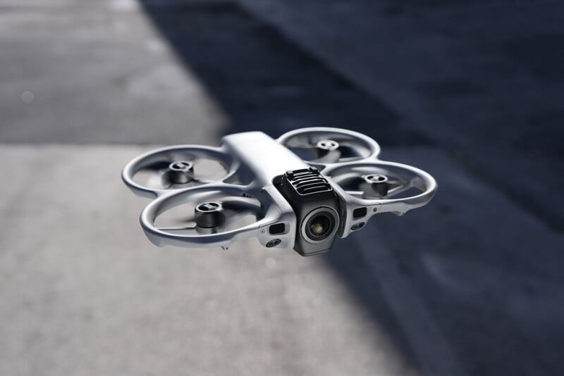 A compact, white quadcopter drone with four propellers hovers close to the ground on a sunlit pavement, casting a shadow. The camera is mounted on the front of the drone.