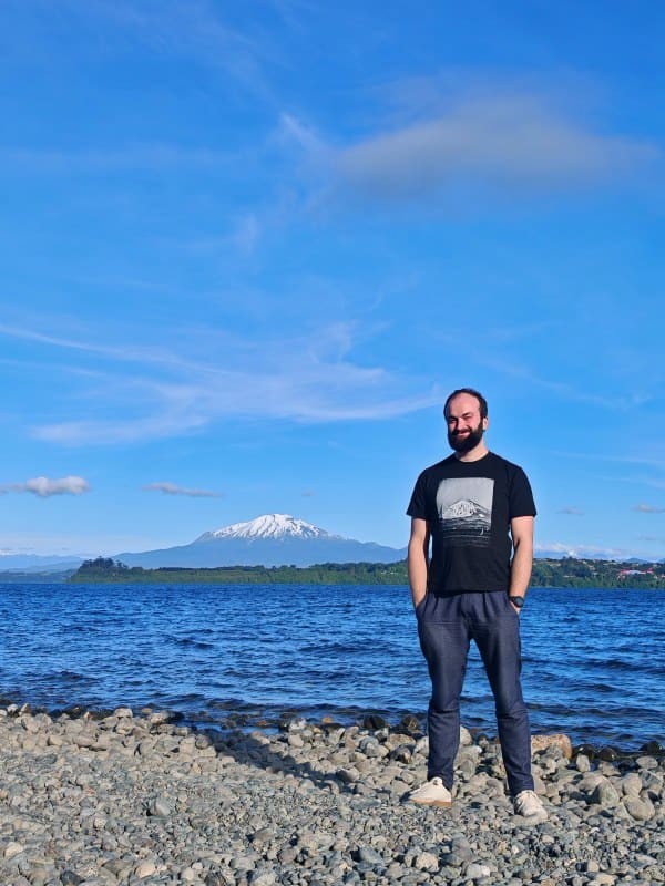 Man standing on rocky shoreline with lake and snow-capped mountain in background.