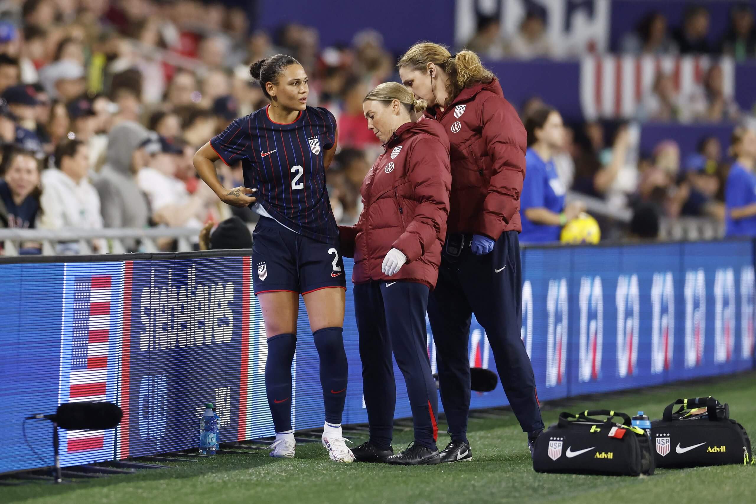 USWNT forward Trinity Rodman, with striped jersey top, is examined by staff members in red jackets.