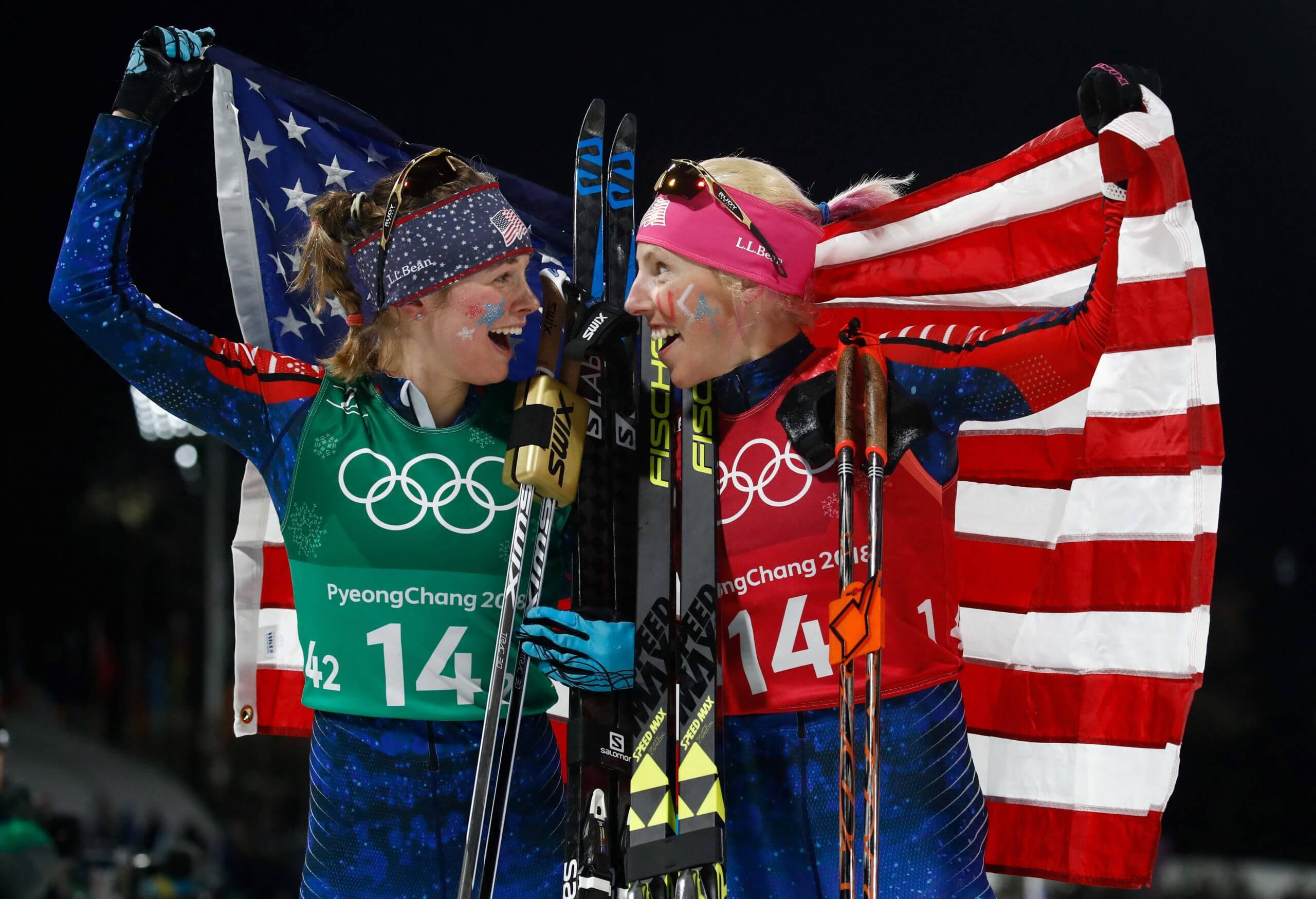 Jessie Diggins and Kikkan Randall stand side by side, looking at each other and smiling, each holding a corner of the American flag draped behind them, after winning gold in the 2018 Olympics.