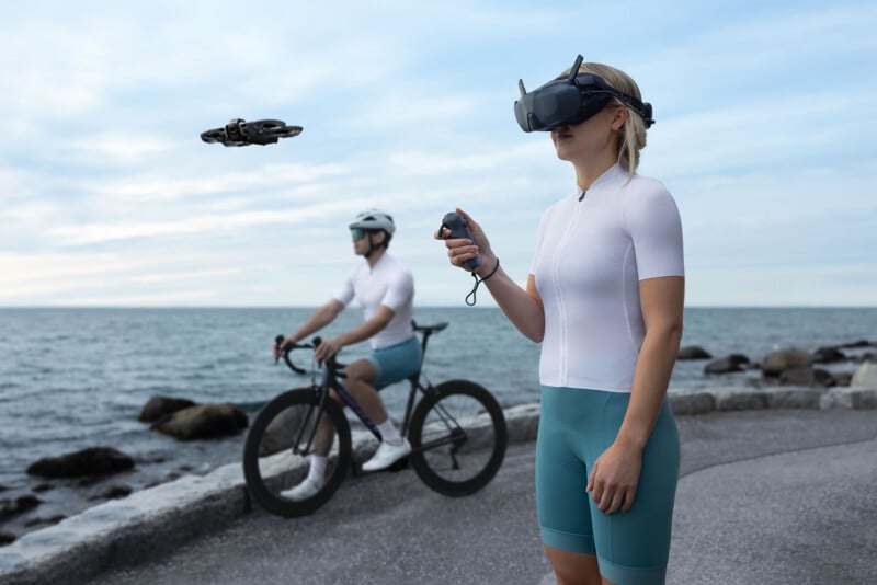 A woman wearing VR goggles and cycling gear operates a flying drone with a remote by the seaside, while a man in similar attire rides a bike in the background on a paved path.