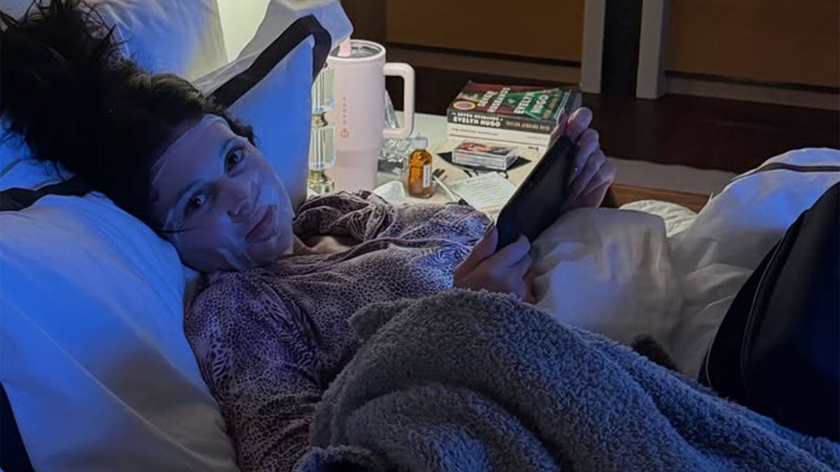 Kylie Jenner in a dark, blue-lit hotel room, wearing animal print pajamas and a white facial sheet mask. She is holding an iPad and smiling broadly at the camera. A table with drinks and books is next to her.