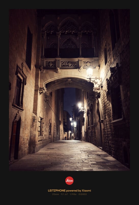 A narrow, cobblestone street at night, illuminated by vintage street lamps, with a decorative stone bridge connecting two old buildings above the alley. Photo taken with a LEITZPHONE powered by Xiaomi.