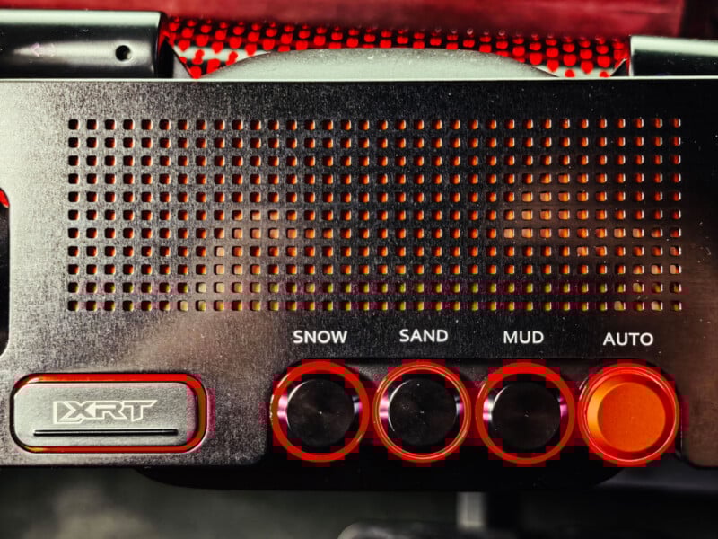 Close-up of a car dashboard panel featuring four red buttons labeled "Snow," "Sand," "Mud," and "Auto," alongside a large button marked "XRT" and a perforated metal grille above the controls.