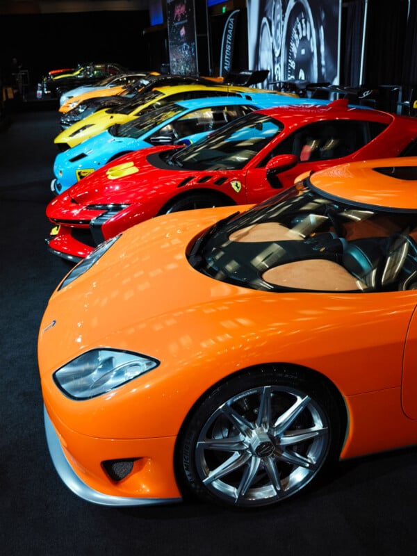 A row of brightly colored exotic sports cars, including orange, red, blue, yellow, and green, are parked indoors on a black carpet at an auto show.