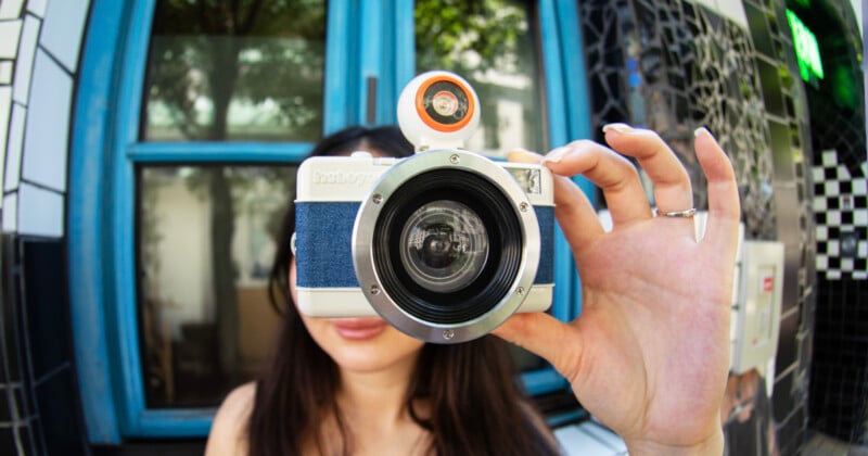 A person holds a large-lens camera up to their face, obscuring their features. The background shows a blue window, green trees reflected in the glass, and a black-and-white tiled wall.