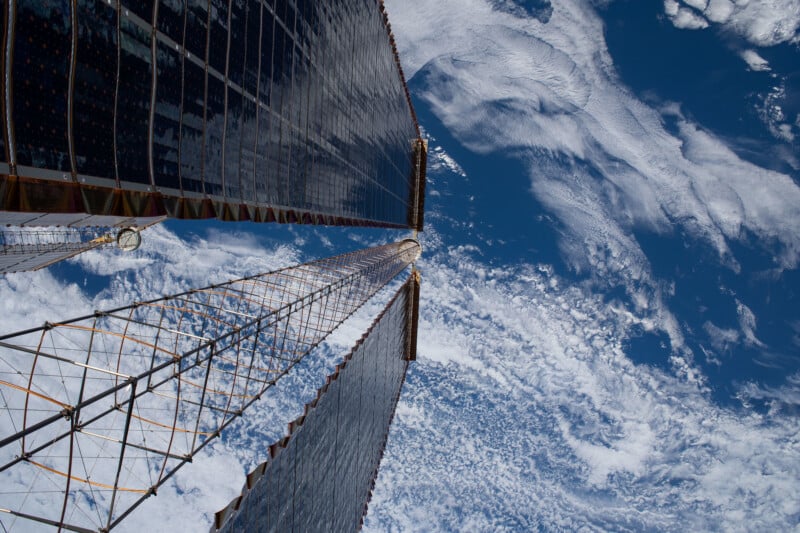Two large solar panels on the International Space Station extend toward Earth, with clouds and blue ocean visible in the background from space.