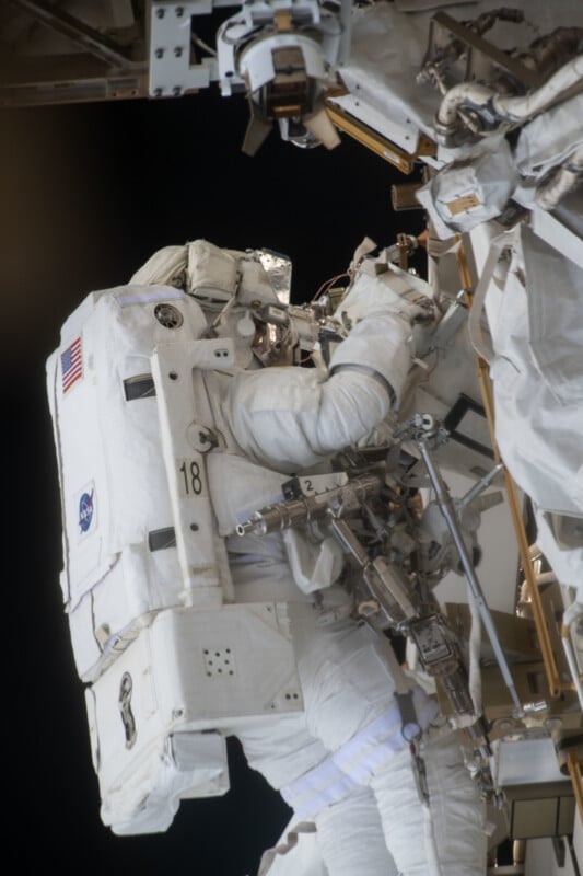 An astronaut in a white spacesuit works on the outside of a spacecraft or space station, surrounded by equipment and wires, with the blackness of space in the background.