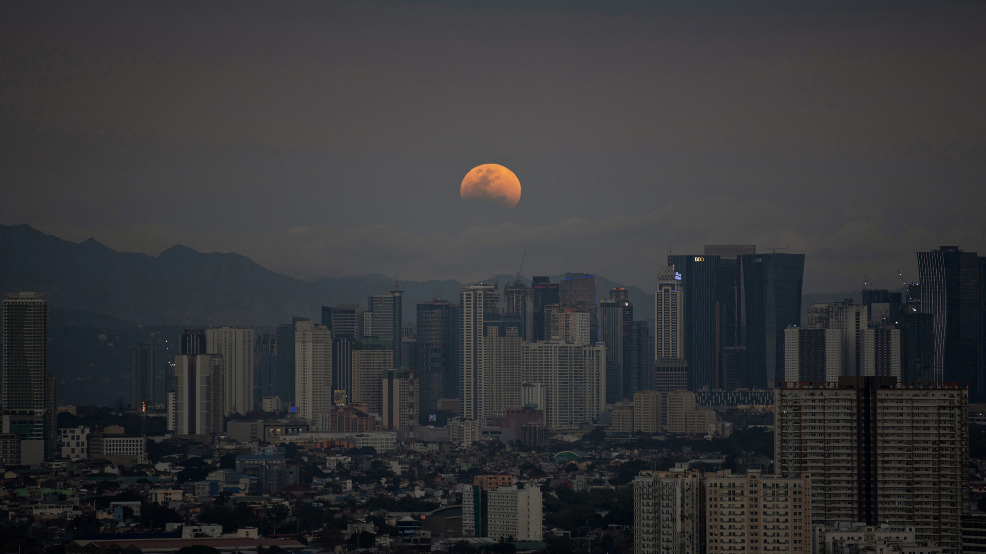 A reddish moon is pictured rising above a city. The lower part of its disk is hidden by Earth's shadow during an eclipse.
