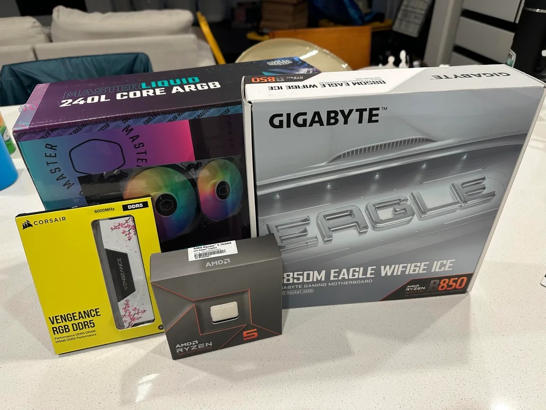 Boxes of computer components including a Gigabyte B850M Eagle WiFi6E Ice motherboard, AMD Ryzen 5 7600X processor, Corsair Vengeance RGB DDR5 RAM, and a Cooler Master Liquid 240L Core ARGB cooler are displayed on a table.
