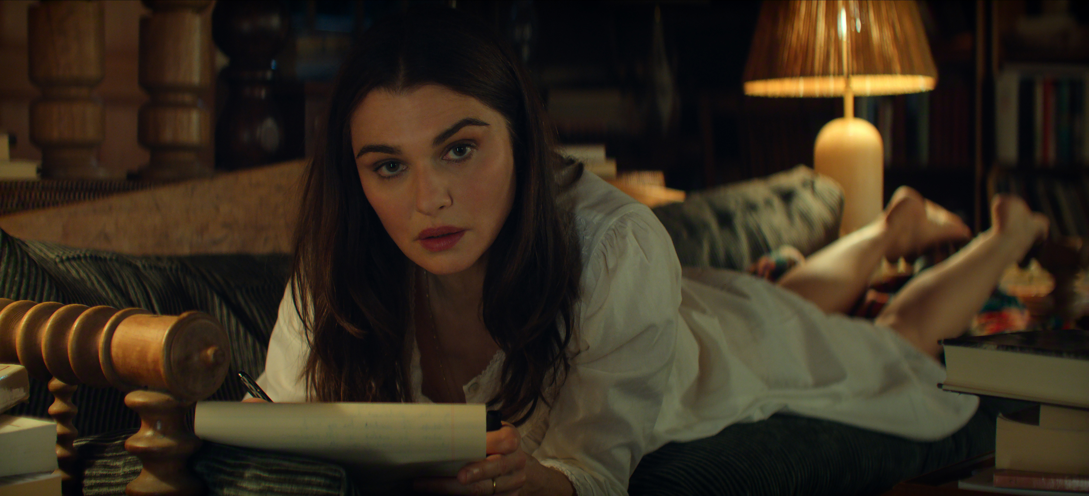 Rachel Weisz as The Protagonist lying on her stomach on a couch writing in a notepad while looking at the camera in Episode 107 of Vladimir