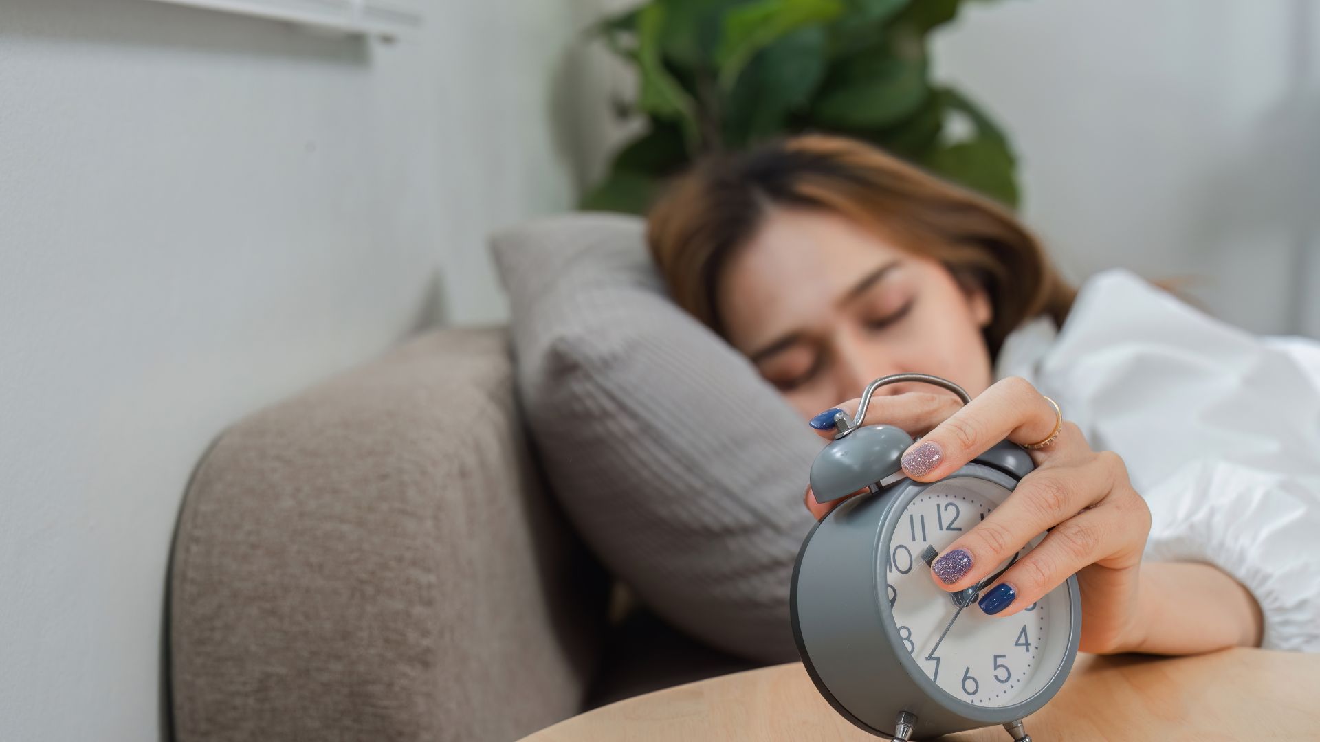 A woman lies in bed and presses snooze on her alarm while her eyes remain closed.
