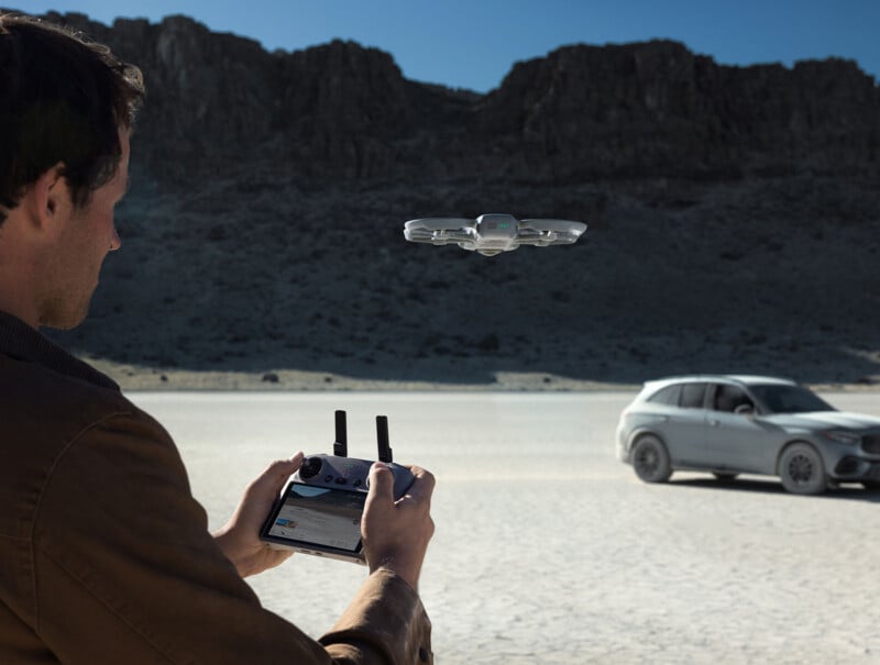 A person operates a drone using a remote controller in a dry, desert landscape with rocky hills in the background and a silver SUV parked on the cracked ground.
