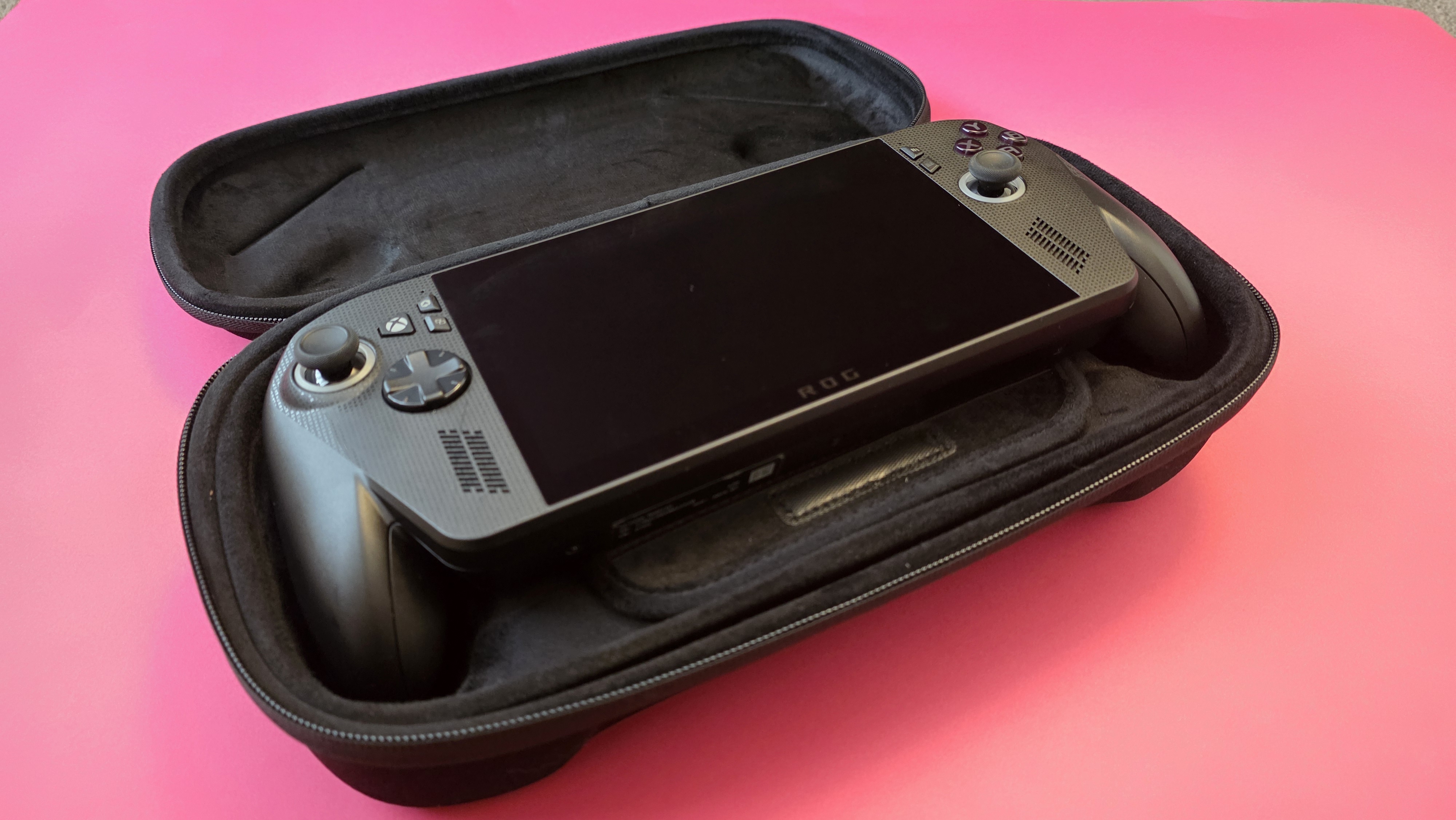 A handheld gaming device with a large screen and controls on each side rests on a pink surface.