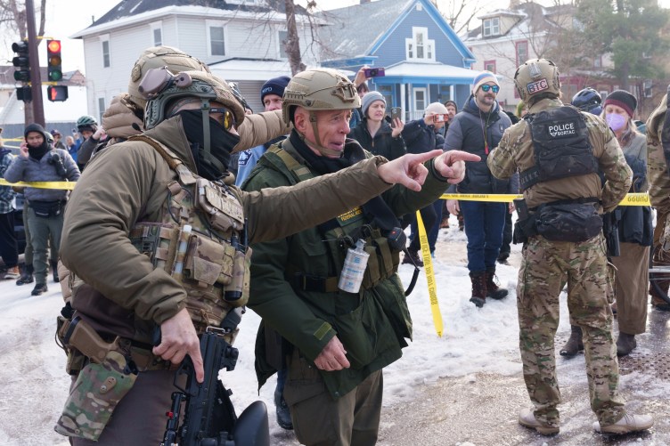 Federal agents point at protesters from inside a yellow-taped perimeter while protesters gather around them in a snowy suburban environment.