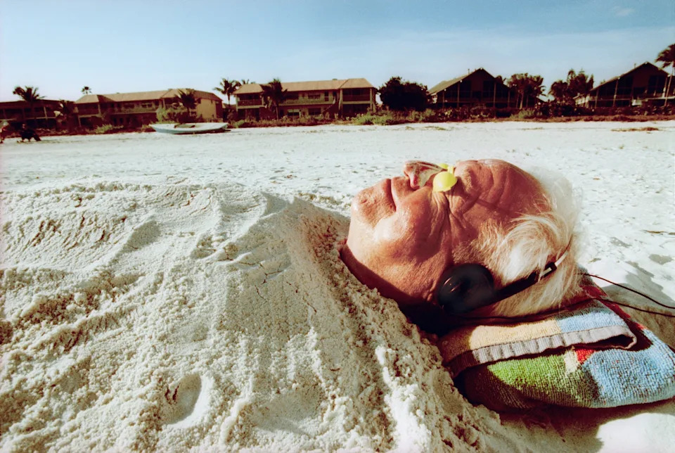 Elderly person on a beach, buried in sand with headphones on, lying on a towel, appears relaxed