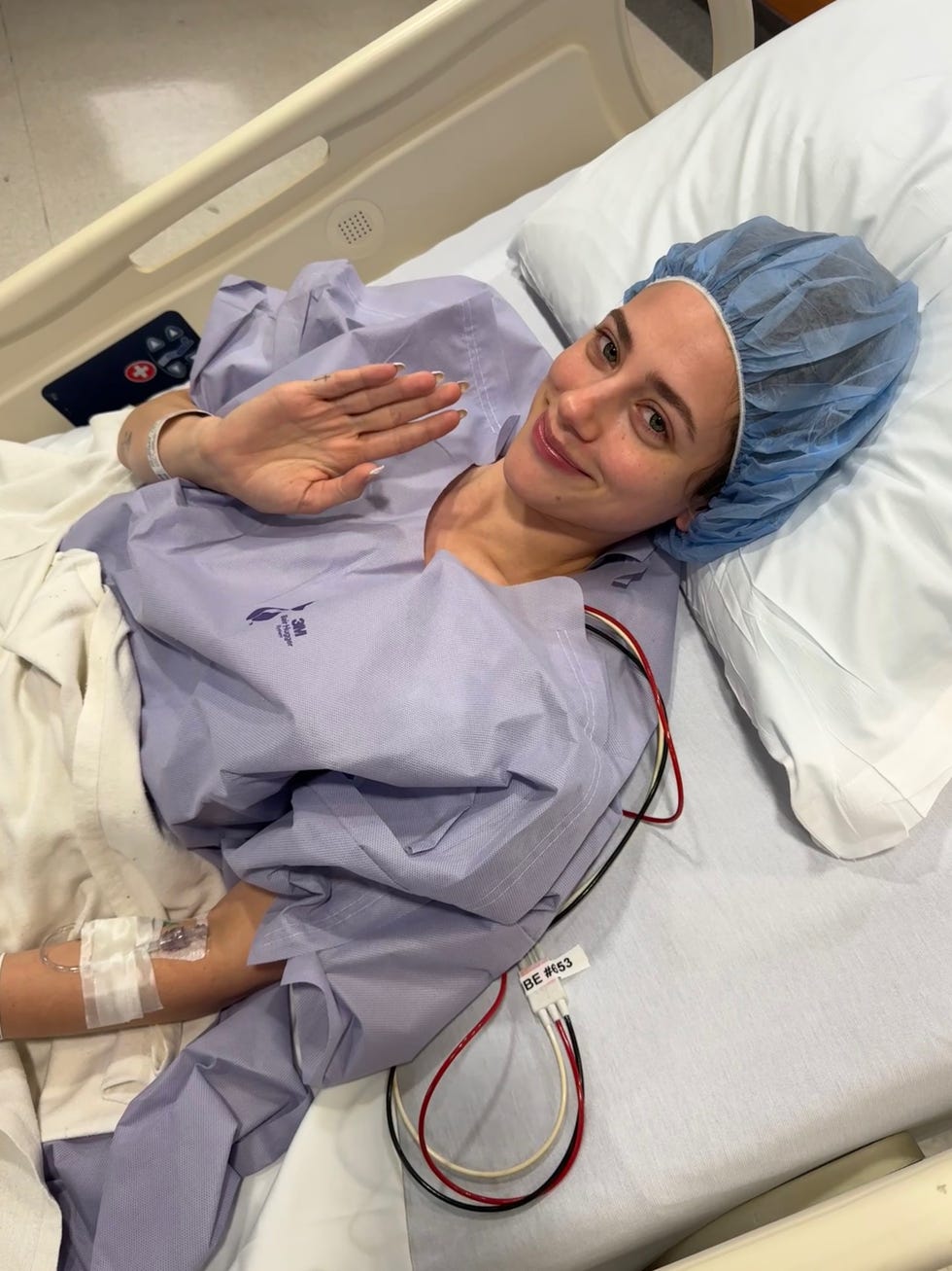 patient in hospital bed wearing a gown and a surgical cap showing a hand gesture