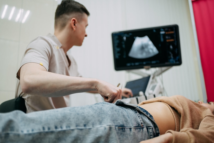 Doctor using ultrasound transducer performing medical examination on female patient's abdomen in hospital.