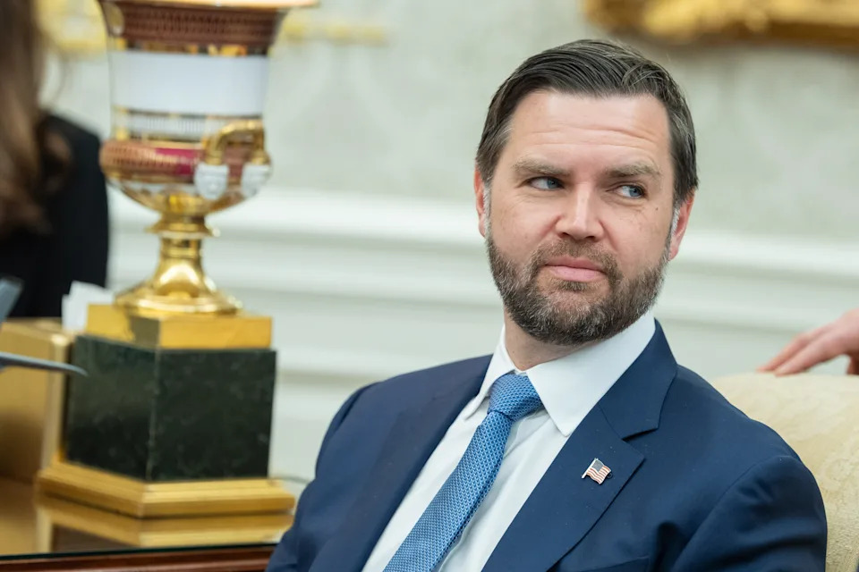 A man in a suit and tie sits in a formal setting, looking slightly to the side. A decorative lamp is in the background