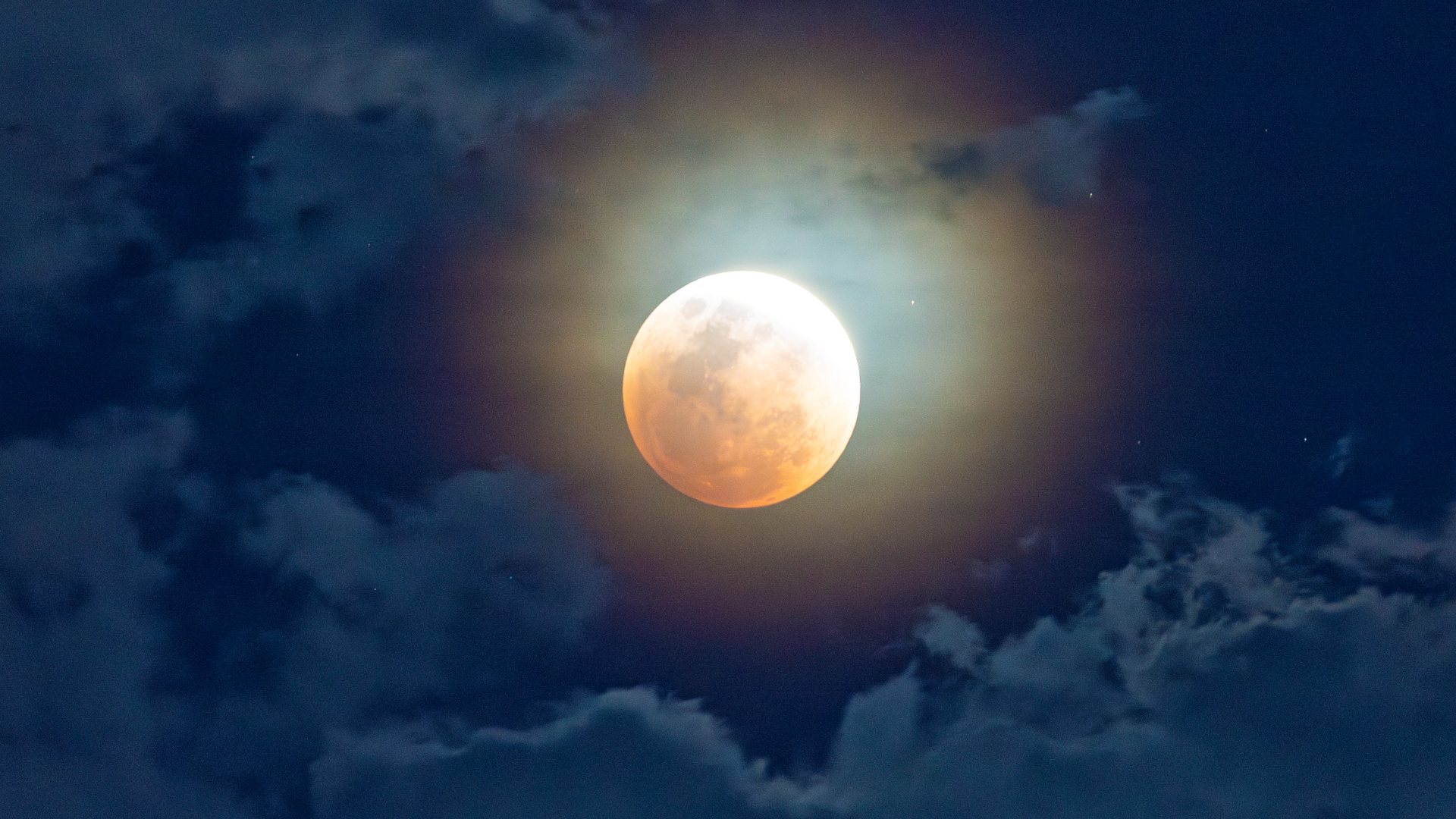A blood moon shines in the night sky between clouds.