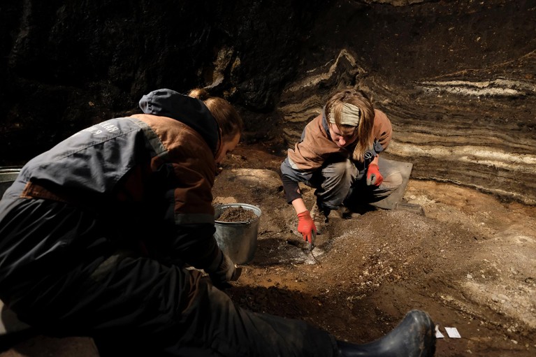 Two archaeologists dig by hand through sediment in a trench.