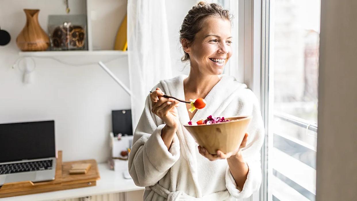 Woman seen smiling, looking out window while eating salad in a robe at home.