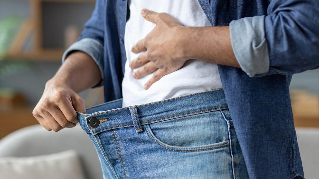 A man shows how much weight he has lost by holding out the waist of his jeans, symbolizing his successful diet.