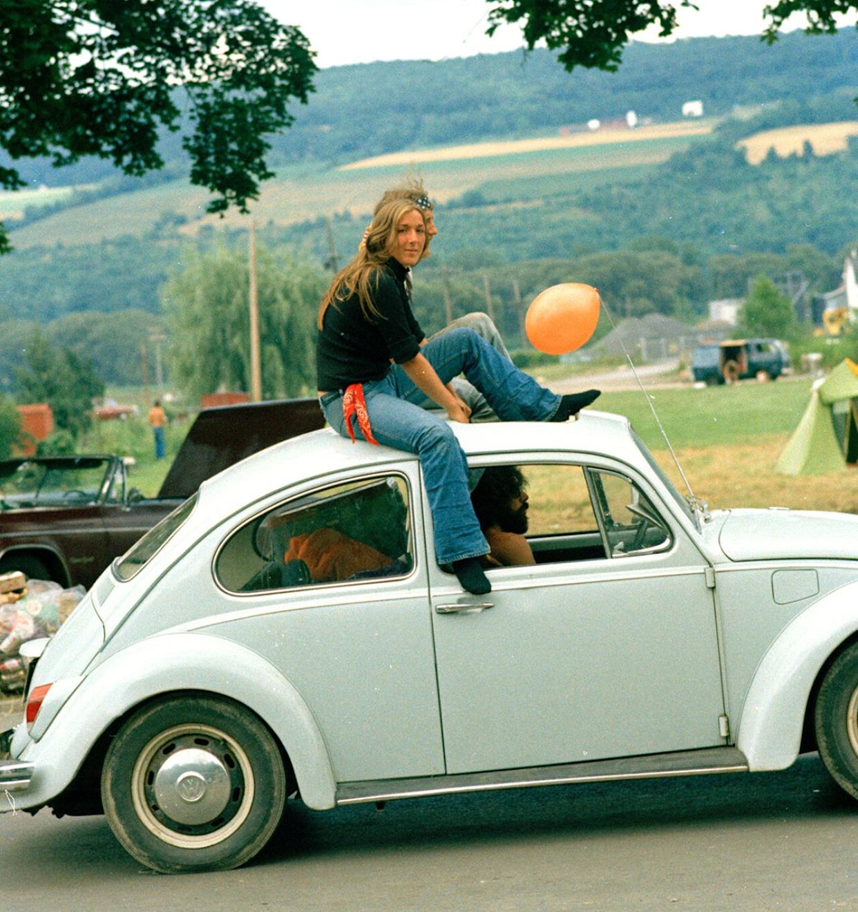 A couple sitting on the roof of a white VW bug driving down the road -