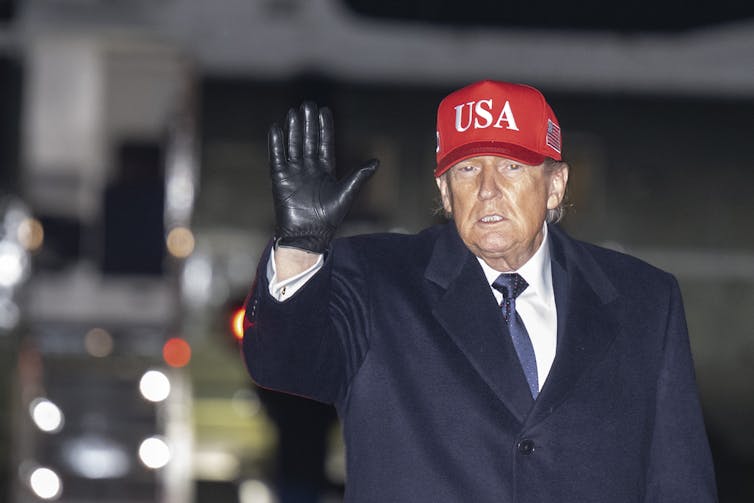 An older man in a blue coat and red hat with 'USA' written on it waves.
