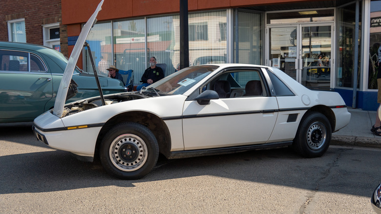 A 1984 Pontiac Fiero with its hood up parked near a building