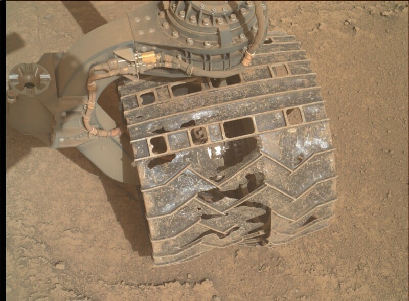 Close-up of a Mars rover's wheel on dusty terrain, showing significant wear with cracks, holes, and scuffed metal on the wheel's surface. Mechanical components and cables are visible above the wheel.